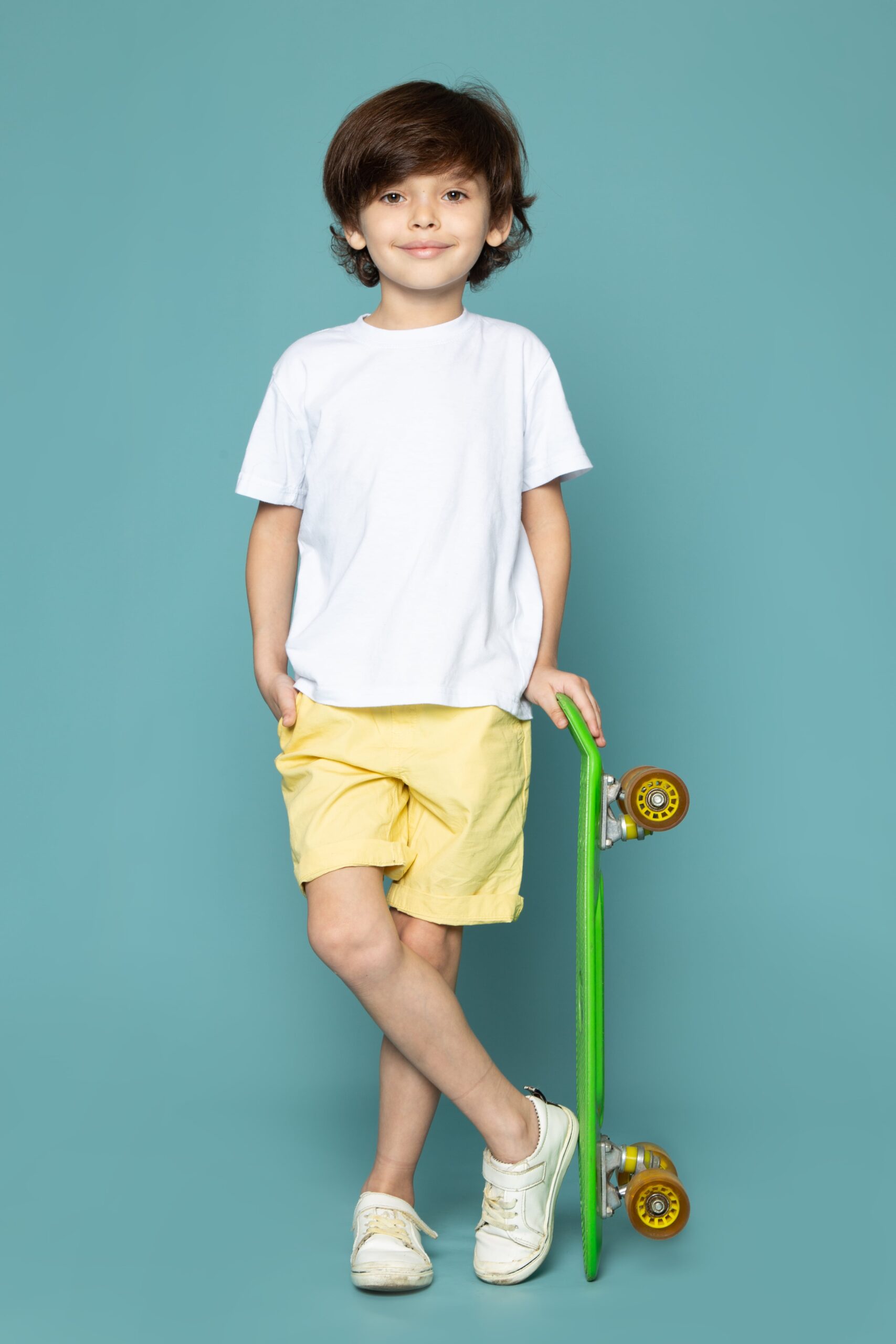 front-view-cute-child-boy-white-t-shirt-yellow-jeans-holding-green-skateboard-blue-floor-min