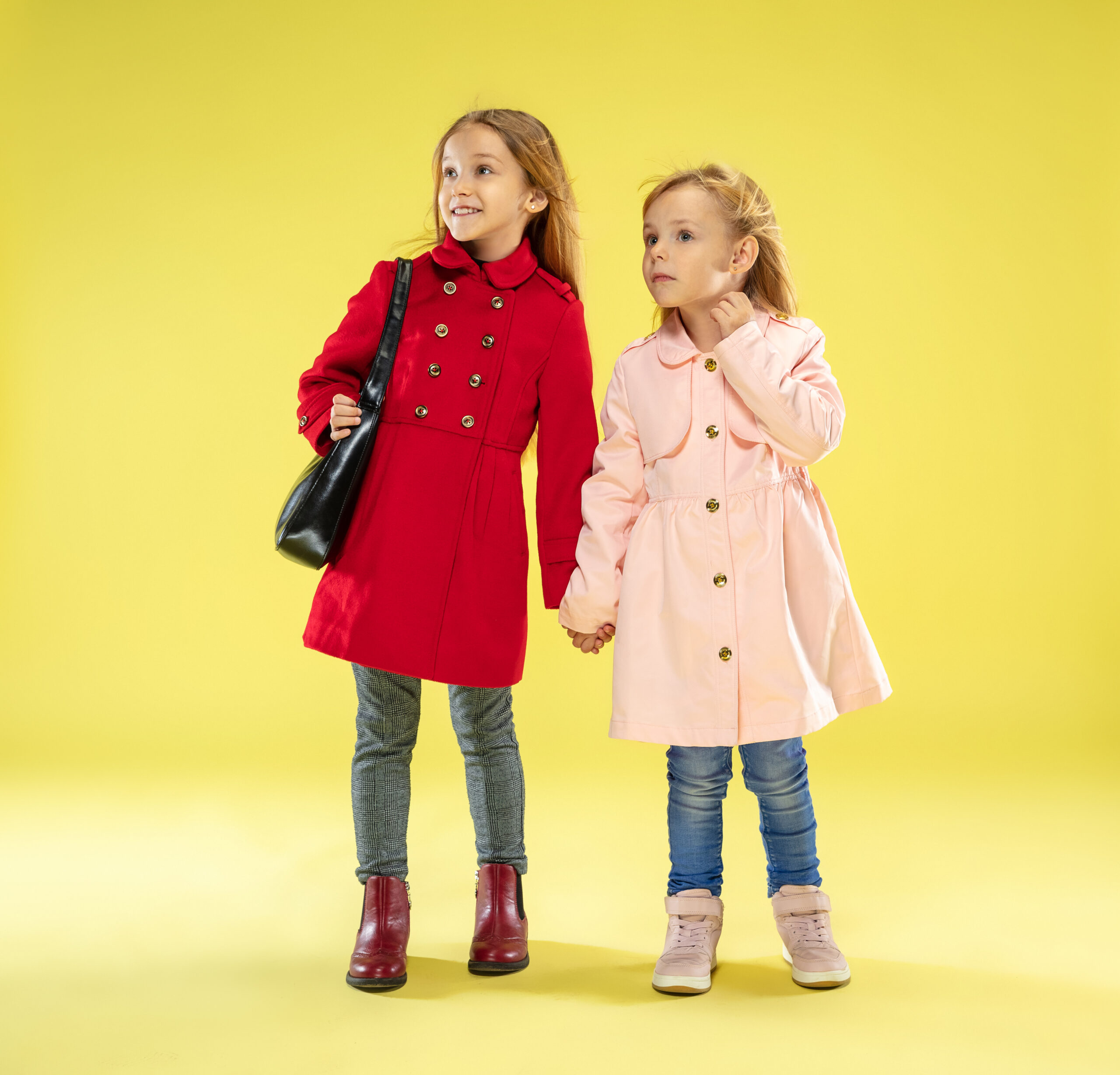 A full length portrait of a bright fashionable caucasian girls in a raincoat holding a black bag on yellow studio background. Autumn and spring fashion for kids. Cute stylish blonde girls.